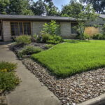 This image depicts the front view of 1300 Cloverleaf Dr. The zoysia grass is lined by river rock near the street and driveway. The yard has several curved beds containing native plants such as yuccas, lantana, cacti, and decorative grasses.