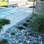 This image depicts a closeup view of the multicolored river rock bed near the front porch. The bed contains several mounded monkey grass, two trailing rosemary plants, and a large Texas Blue stem grass.
