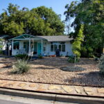 A street image of 1424 Corona Dr. with a yard consisting of river rock, native plants. and a flagstone pathway leading to the front Turquoise door.