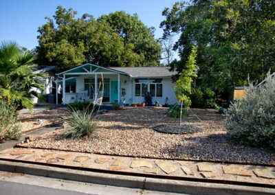 A street image of 1424 Corona Dr. with a yard consisting of river rock, native plants. and a flagstone pathway leading to the front Turquoise door.