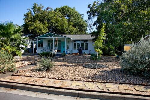 A street image of 1424 Corona Dr. with a yard consisting of river rock, native plants. and a flagstone pathway leading to the front Turquoise door.