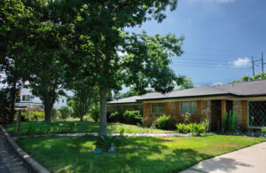 A front view of 5703 Sutherlin with a shady yard and red brick home and a red stone pathway leading to the front door.