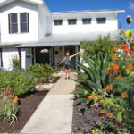 This image shows the owner of 5400 Wellington standing proudly amongst her beautiful front yard garden. You'll find a large blue agave, several blue salvias, canna lilies infant of the porch, and more colorful Pride of Barbados.