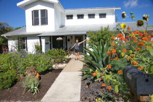 This image shows the owner of 5400 Wellington standing proudly amongst her beautiful front yard garden. You'll find a large blue agave, several blue salvias, canna lilies infant of the porch, and more colorful Pride of Barbados.
