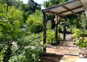 This image depicts the overhang structure added on to the front porch. It is has a metal cover and natural wood columns. In front of the porch is a cluster of small red cannas. hanging from the structure is a flower pot full of purslane and a small wind chime.