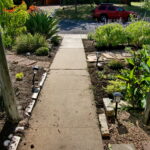 An image taken from the perspective of the front porch looking out into the yard. It shows the sidewalk leading to the street with beds of agave, salvia, cannas, and yellow daisies lining the sidewalk.